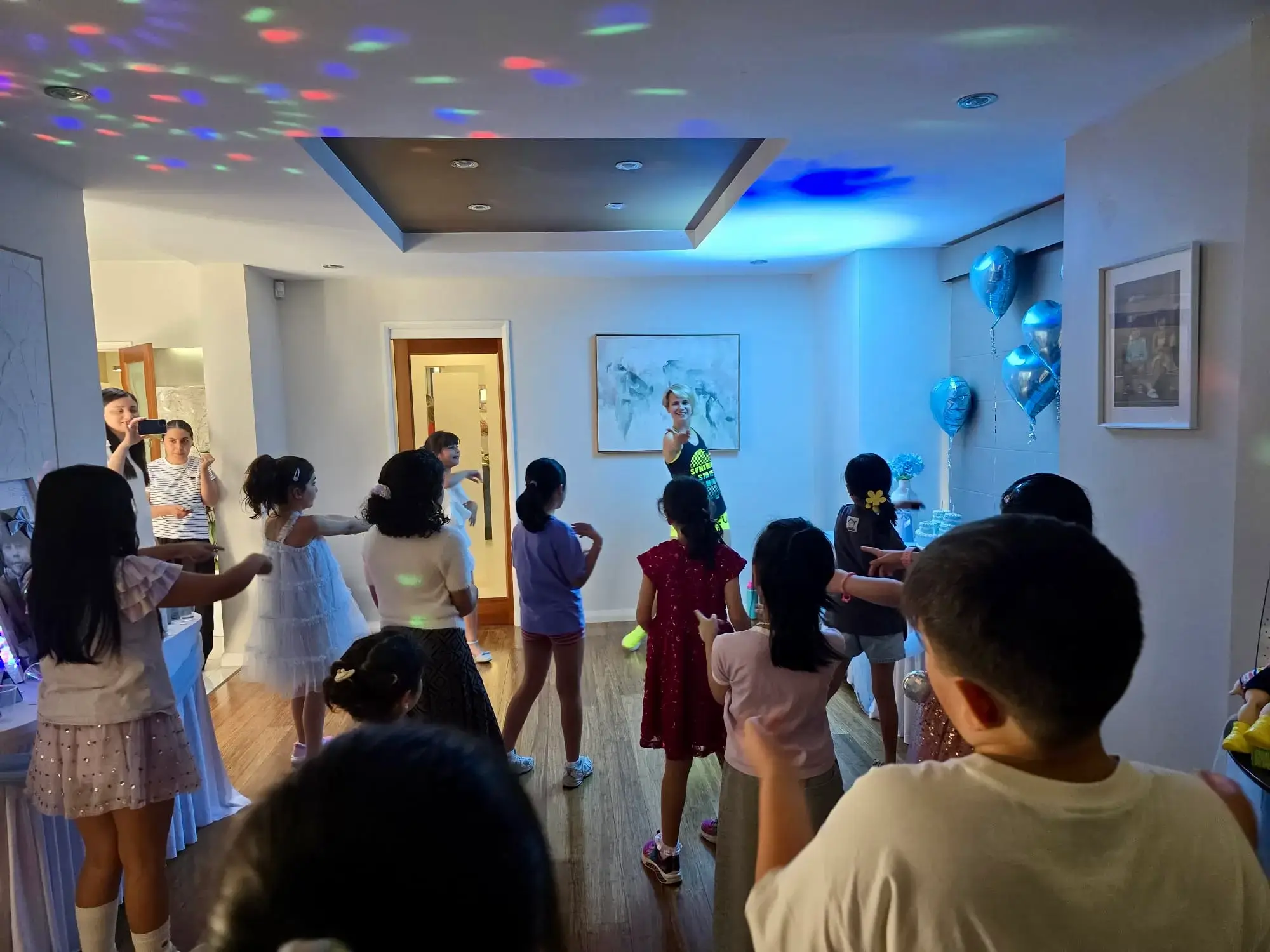 Professional dance party entertainer leading a room of kids in a dance routine under disco lights, with blue balloons on the wall