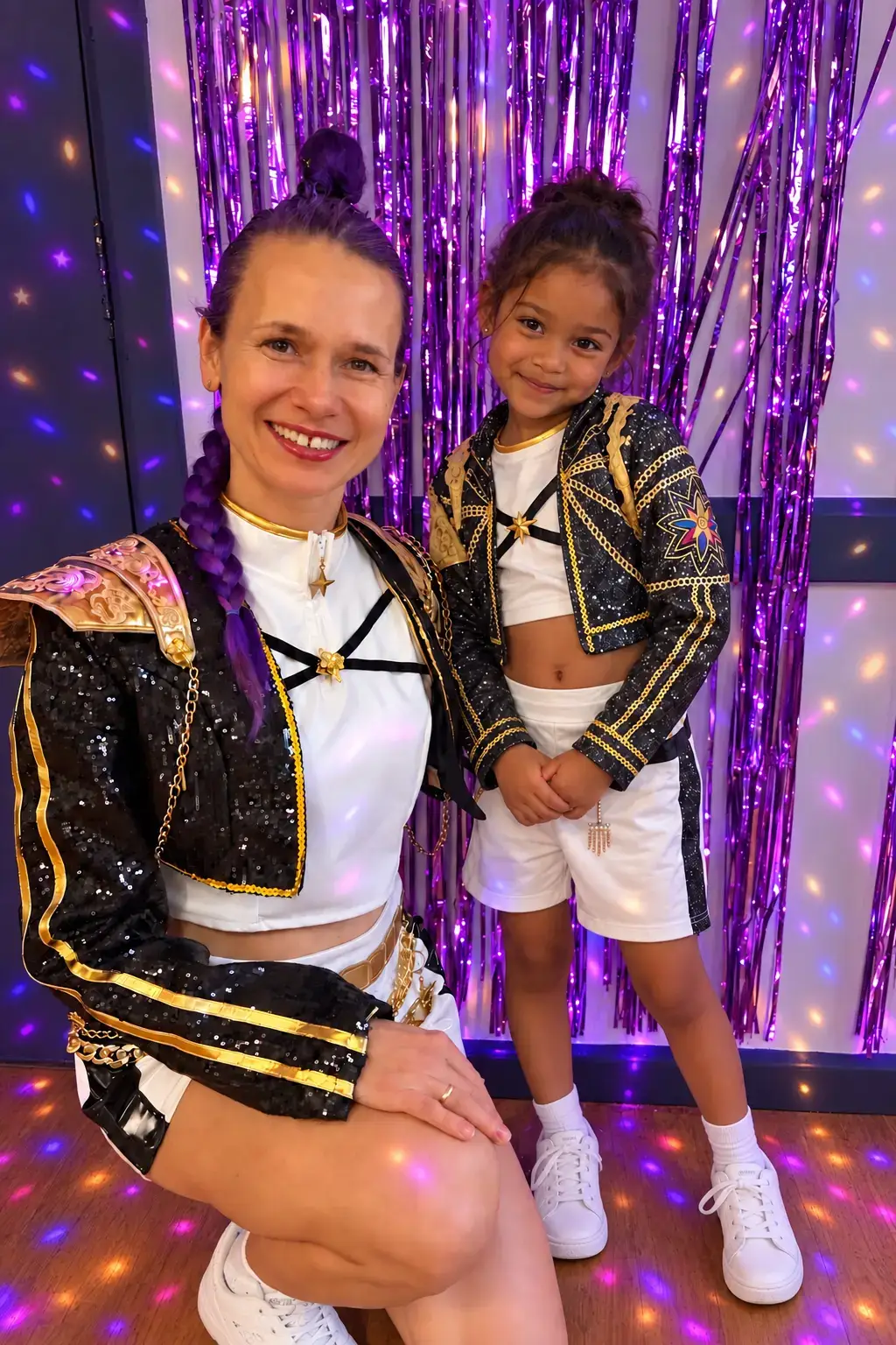 Elena and a girl in matching black and gold K-Pop jackets smiling in front of a purple tinsel backdrop
