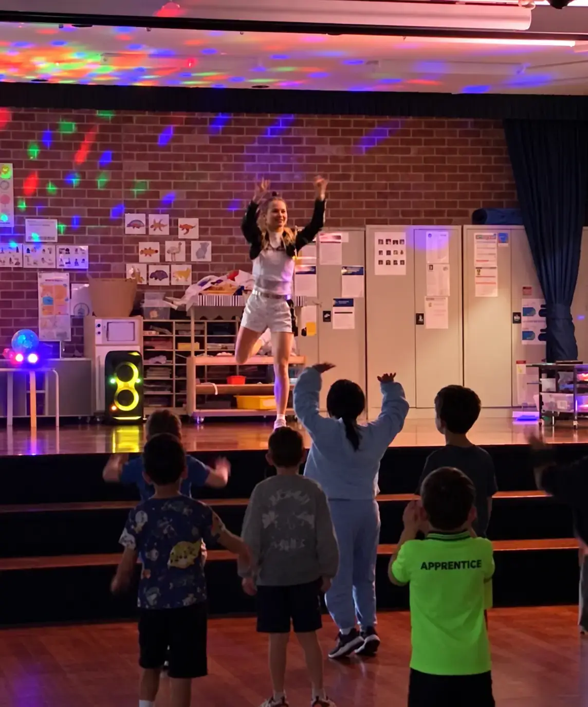 Professional dance party entertainer in K-Pop costume leading kids from the stage of a hall under disco lights
